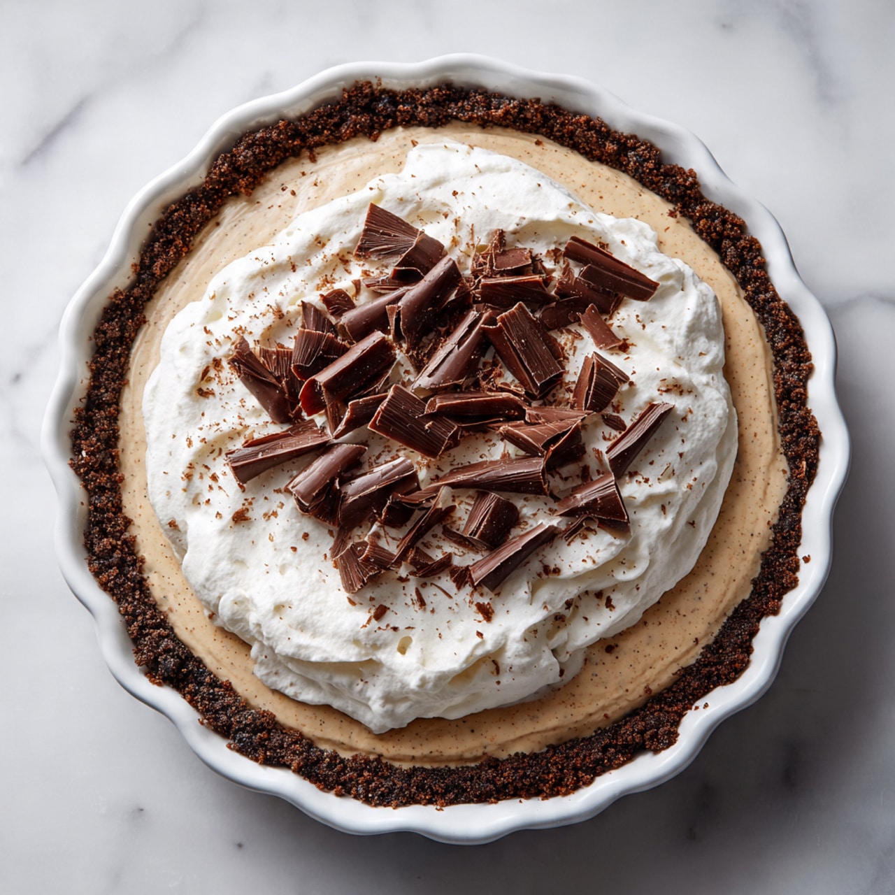 A close-up of a three-layered dessert slice on a white plate with a white marbled texture underneath. The bottom layer is dark brown and crumbly, resembling a chocolate cookie crust. The middle layer is thick, light brown, and airy with a mousse-like texture. The top layer is smooth, thick, and white, looking like whipped cream with soft peaks along the edges. On top, there are two curled dark chocolate shavings, one larger and one smaller, adding a decorative touch. Some small crumbs are scattered on the plate around the dessert. photo taken with an iphone --ar 4:5 --v 7