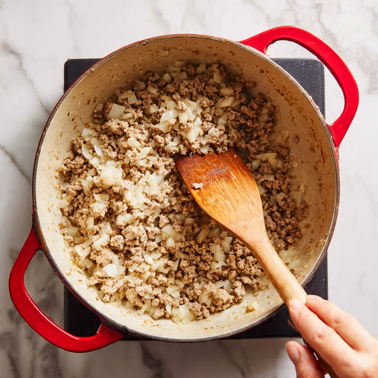 A close-up view of a red pot on a black stove top, filled with a mix of cooked ground meat and finely chopped onions. The inside of the pot is cream-colored, slightly stained from cooking. A wooden spatula stirs the mixture, held by a woman's hand, showing the browned texture of the meat with softer, lighter pieces of onion mixed throughout. The background features a white marbled texture. photo taken with an iphone --ar 4:5 --v 7