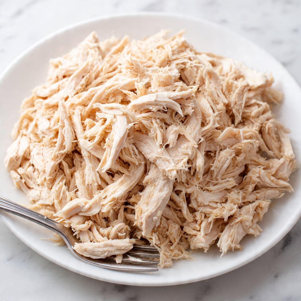 The image shows a large pile of shredded cooked chicken pieces that are light beige in color and have a soft, fibrous texture. The chicken fills almost the entire visible area and is spread evenly on a white plate. A silver fork is partially visible at the bottom left corner of the image, touching some of the shredded chicken. The background is a white marbled surface, giving a clean and simple look. photo taken with an iphone --ar 4:5 --v 7