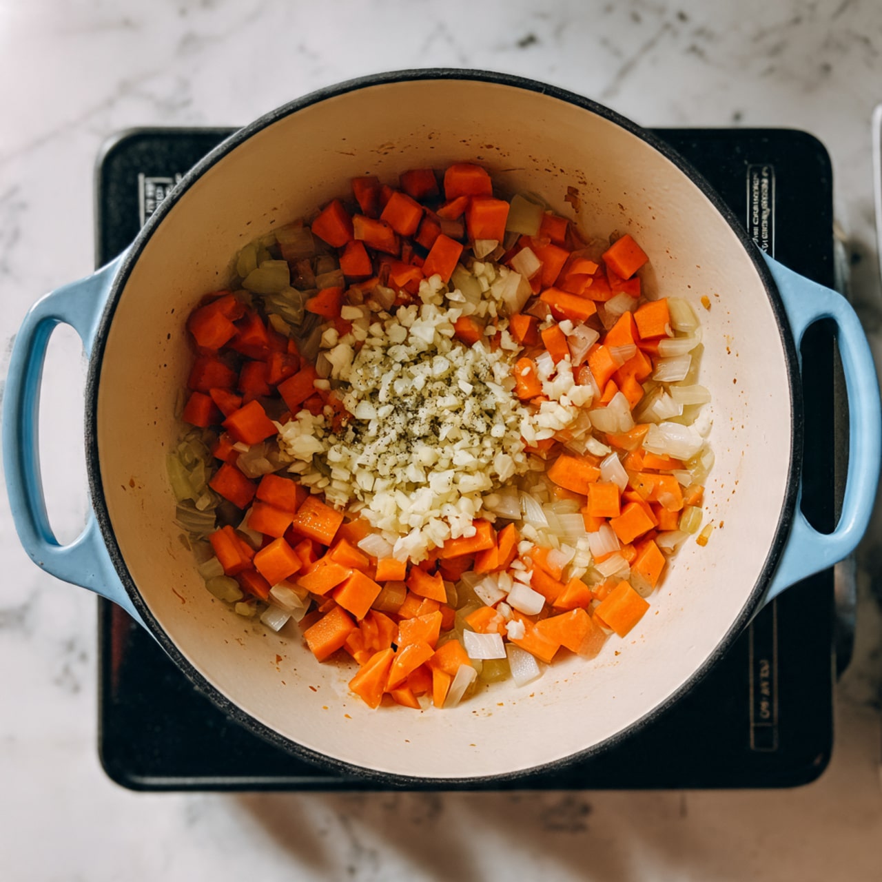 A close-up top view of a white cast iron pot with light blue handles on a black electric stove, placed on a white marbled surface. Inside the pot, there is a mix of diced orange carrots and translucent cooked onions forming the bottom and middle layers, surrounding a small pile of finely chopped white garlic in the center. The texture of the vegetables looks soft and cooked, with some brown bits sticking lightly to the pot's bottom. photo taken with an iphone --ar 4:5 --v 7