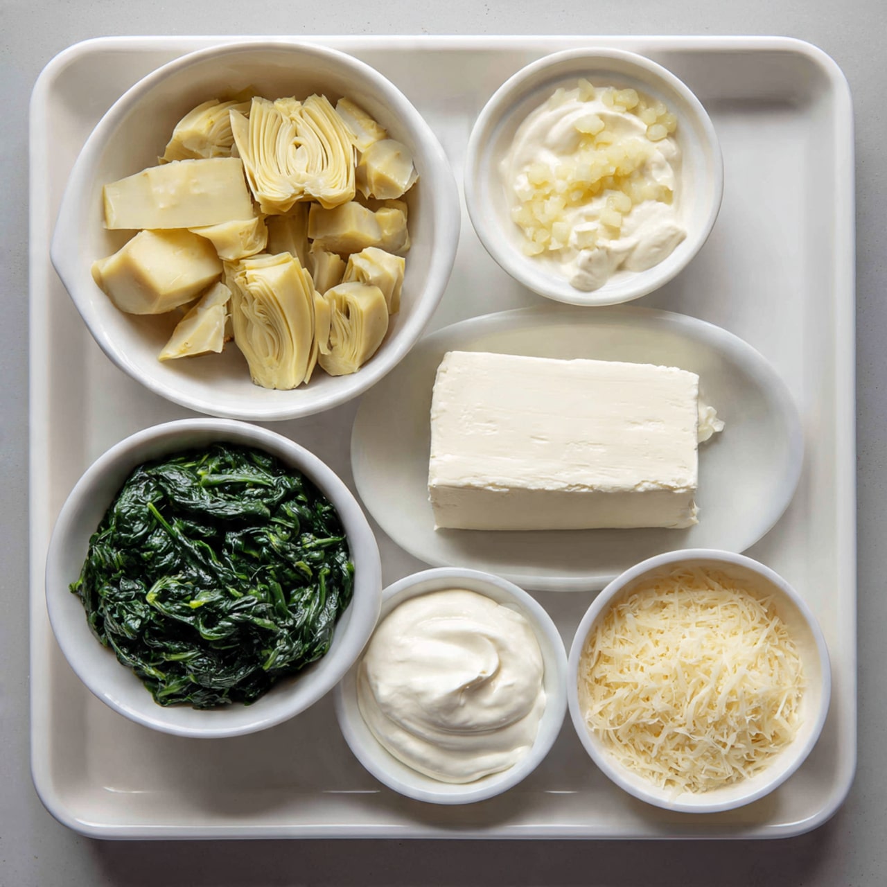 The image shows a white tray with seven separate white bowls and plates, each holding different ingredients for a dish. In the top left bowl, there are pale yellow artichoke hearts cut into pieces, sitting loosely. Next to it on the right is a small bowl with creamy white mayonnaise. Below the mayonnaise, there is a tiny bowl filled with finely chopped pale garlic pieces. To the right of the garlic, a flat white plate holds a thick block of smooth white cream cheese with soft edges. In the bottom row, starting from the left, a small bowl contains thick, white sour cream with a smooth texture. In the center bottom bowl, there is a generous amount of dark green frozen spinach, loosely piled with a slightly rough leafy texture. Finally, on the right bottom side, a small bowl holds finely grated pale yellow Parmesan cheese. The white tray rests on a light gray surface. Photo taken with an iphone --ar 4:5 --v 7