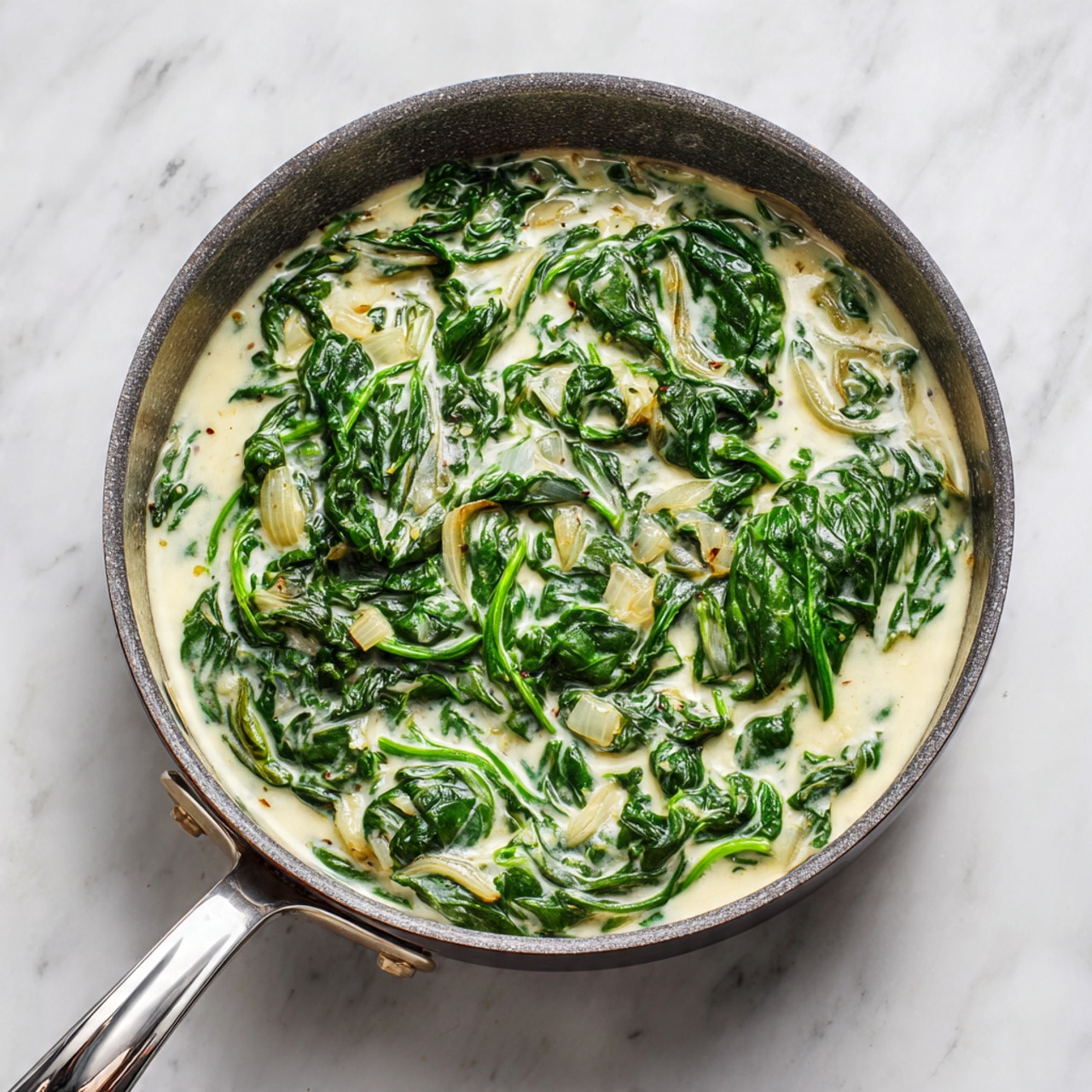 A gray pan filled with creamy cooked spinach mixed with cream sauce and small pieces of soft onions, the dark green wilted spinach leaves spread evenly throughout the light-colored creamy sauce giving a rich, smooth texture. The pan is placed on a white marbled surface, with the metal handle shown clearly at the bottom. Photo taken with an iphone --ar 4:5 --v 7