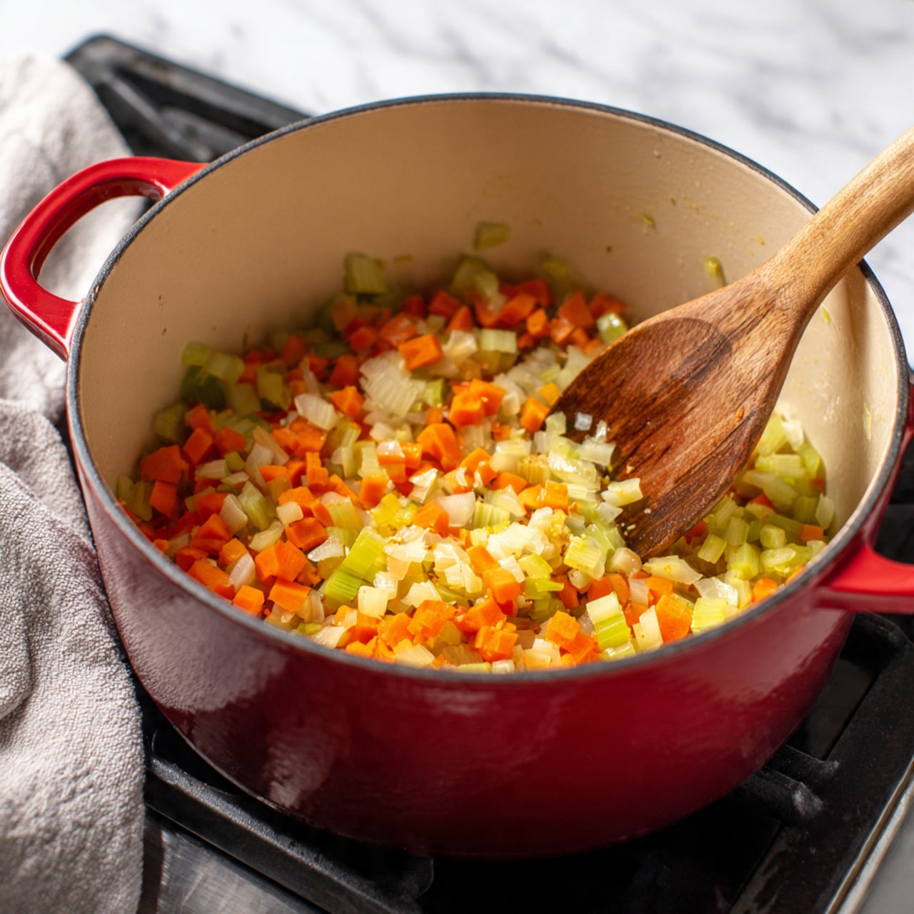 The image shows a red pot with a light beige inside filled with diced vegetables being cooked. The vegetables include bright orange carrots, light green celery, and small bits of translucent onions. A wooden spoon with a smooth brown texture is stirring the vegetables on the right side of the pot. The pot is placed on a stove with a shiny black surface, and a soft gray cloth is seen casually laid in the background over a white marbled surface. The scene is bright and clean, highlighting the fresh colors of the vegetables. photo taken with an iphone --ar 4:5 --v 7