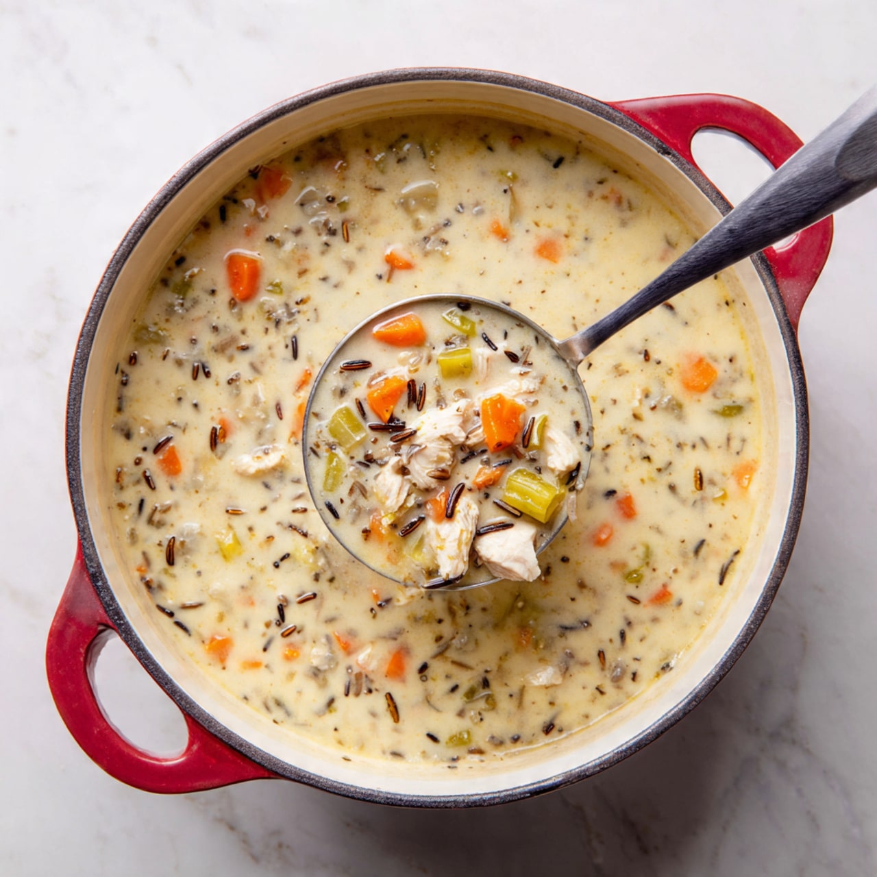 A large white pot with a red outer edge is filled with creamy soup. The soup has three main layers: a thick white creamy base, small orange carrot pieces scattered throughout, and small green celery chunks. There are also bits of white chicken and black wild rice mixed in. A large metal ladle holds up a spoonful of the soup, showing the small chunky ingredients clearly. The pot sits on a white marbled surface. Photo taken with an iphone --ar 4:5 --v 7