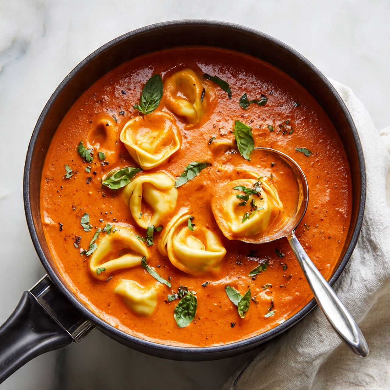 A white bowl filled with a creamy orange tomato soup, in which soft, folded tortellini pasta float evenly throughout. The soup has a smooth texture with small dotted specks of black pepper on top. Fresh dark green basil pieces are scattered across the surface, adding color contrast. The bowl sits on a white marbled surface, showing part of the background. photo taken with an iphone --ar 4:5 --v 7