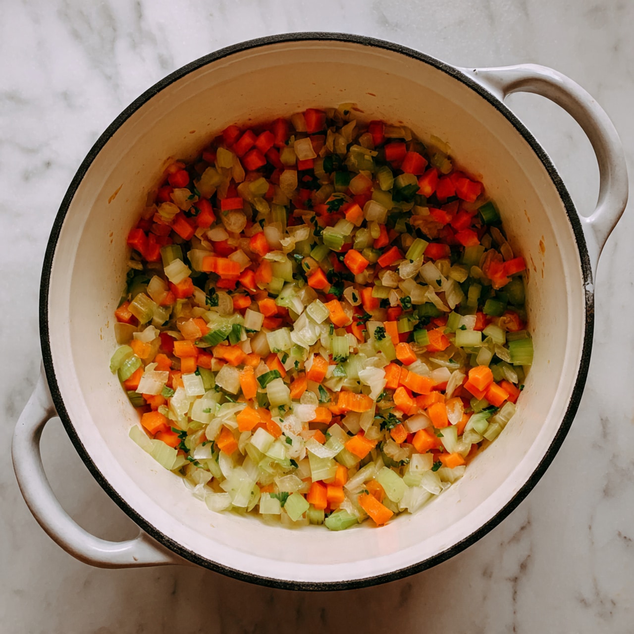A white cooking pot with dark rim and handles, filled with small pieces of sautéed vegetables. The vegetables are mainly three layers: diced orange carrots, light green celery pieces, and chopped translucent onions with a soft texture. They are mixed evenly inside the pot showing a colorful mix. The pot is placed on a surface with white marbled texture. Photo taken with an iphone --ar 4:5 --v 7