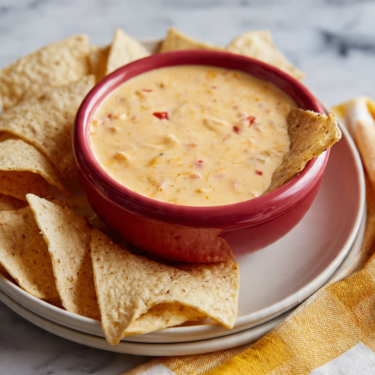 A red bowl filled with a creamy light orange dip that has visible chunks of tomatoes, placed on a black plate. Around the bowl, there are several light beige tortilla chips arranged casually. The plate is set on a yellow cloth with thin white stripes, and the whole scene is on a white marbled surface. photo taken with an iphone --ar 4:5 --v 7