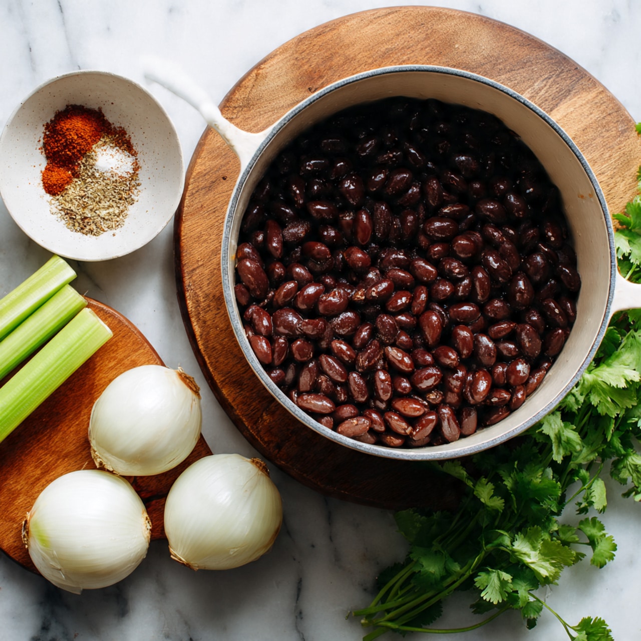 A white pot filled with a single layer of shiny dark brown beans sits on a round wooden board on a white marbled surface. Around the pot are three celery sticks on the left, two halved white onions at the bottom, and a small white bowl at the top left filled with a mix of light brown, crushed red, and white spices. On the right side, a close-up of fresh green cilantro leaves with visible veins rests on the same white marbled surface. photo taken with an iphone --ar 4:5 --v 7
