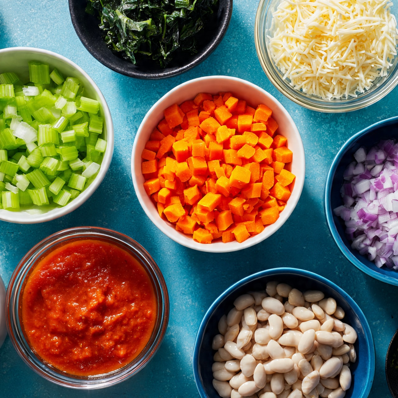The image shows seven bowls and a small glass of ingredients arranged on a blue surface. There are two white bowls holding bright chopped celery and vibrant orange diced carrots, while three blue bowls contain pale white beans, finely chopped purple onions, and minced garlic, each showing clear textures. A small transparent container holds pale yellow shredded cheese and a clear glass cup is filled with smooth red tomato sauce. In the corner, there is a black bowl with dark green leafy vegetables. All items are neatly spaced and the colors are vivid and fresh. Photo taken with an iphone --ar 4:5 --v 7