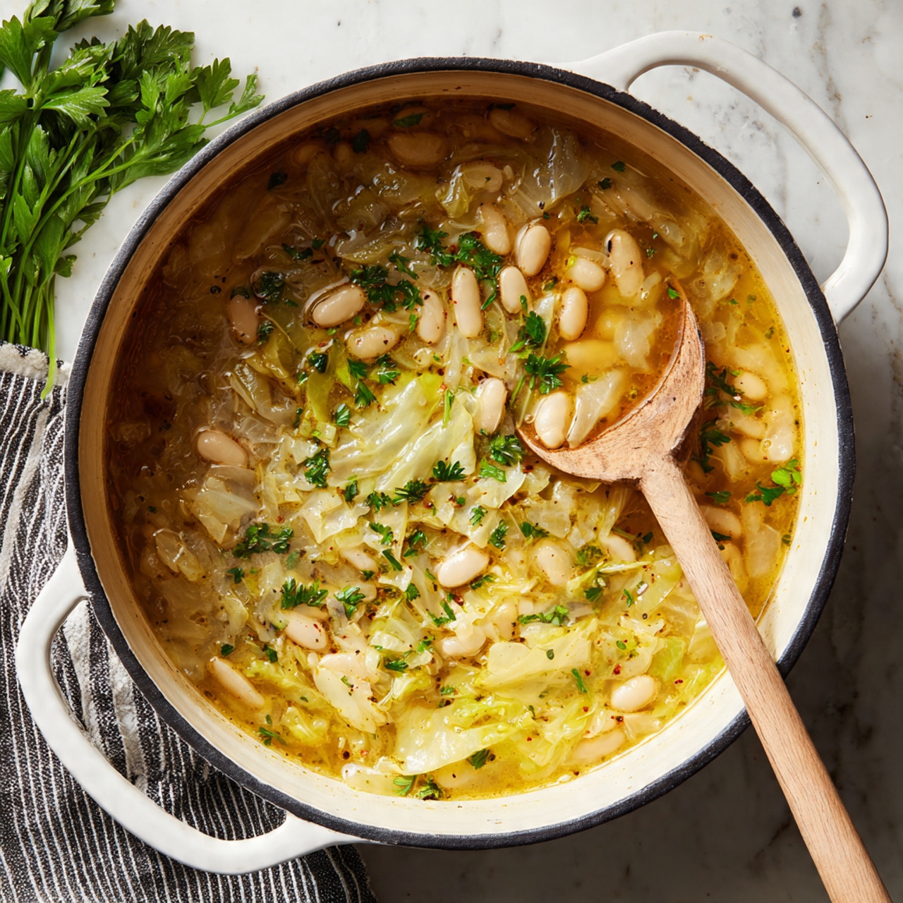 A round white cast iron pot with a black inside edge holds a thick soup with layers showing clear white beans on the top left side, and a golden-yellow broth with cooked cabbage and spices on the right side, sprinkled with small green herb leaves. A wooden spoon rests inside the pot, dipped in the broth near the bottom right. The pot is placed on a white marbled surface, next to a black and white striped cloth and a small bunch of fresh green herbs on the left side. Photo taken with an iphone --ar 4:5 --v 7