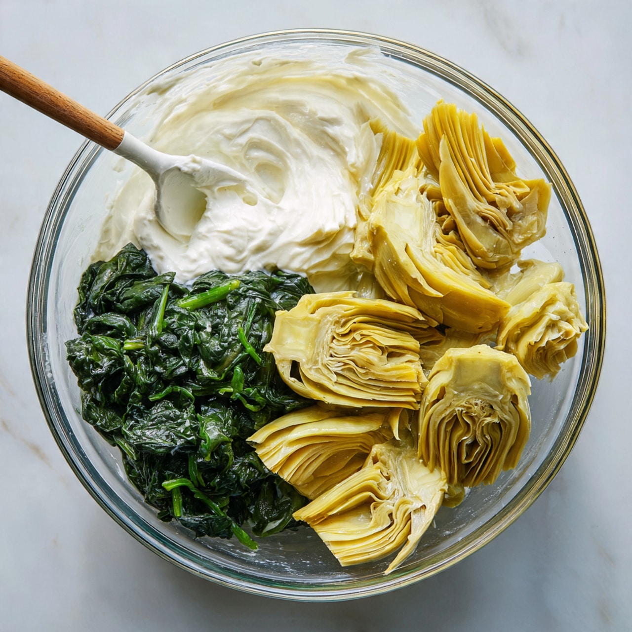 A clear glass bowl sits on a white marbled texture, containing three distinct layers of ingredients. On the left, there is a white creamy mixture with a smooth texture, next to it on the right is a pile of light yellow artichoke pieces with a soft, folded look. At the bottom left of the bowl, deep green cooked spinach with a slightly wet and leafy texture is present. A white spatula rests inside the bowl on the left side, partially covered with the white creamy mixture. photo taken with an iphone --ar 4:5 --v 7