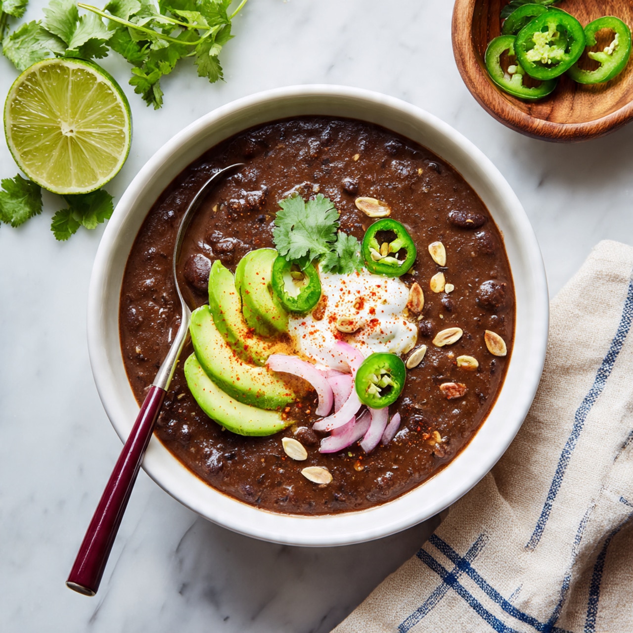 A white bowl filled with smooth dark brown black bean soup. On top, there are three light green slices of avocado placed in a row on the right side, some bright green sliced jalapeño peppers scattered around, a dollop of white sour cream near the center with a sprinkle of red chili powder, thin rings of pink pickled onions to the right of the sour cream, and a few green cilantro leaves and light brown pumpkin seeds spread on top. A spoon with a dark red handle is partially dipped into the soup from the left side. The bowl is set on a white marbled surface with a halved lime above, a small wooden bowl with more jalapeño slices to the top right, and a bunch of cilantro partly visible on the left. A cream cloth with blue stripes is placed near the bowl's bottom right corner. photo taken with an iphone --ar 4:5 --v 7