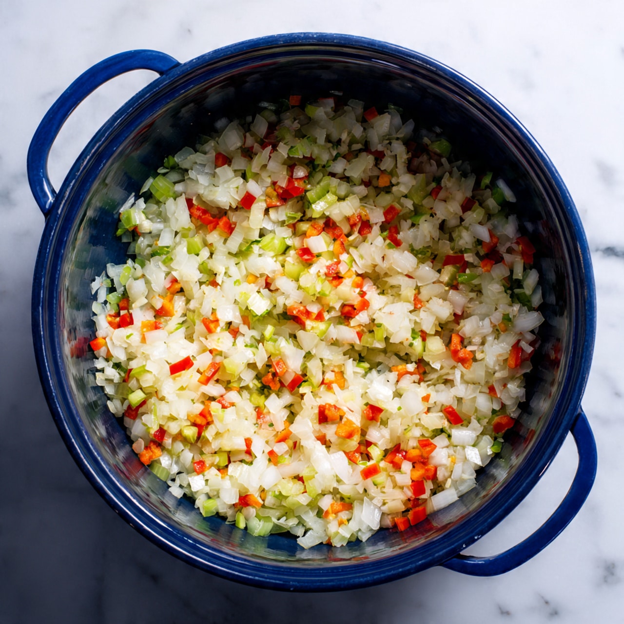 A large dark blue pot sits on a stove with a white marbled surface underneath, filled with finely chopped vegetables. The vegetables form a colorful mix in the pot’s single visible layer, mainly white onions mixed with small pieces of red bell peppers and light green celery. The texture looks soft but still slightly firm, showing that the vegetables are cooking but not fully softened yet. The pot has two blue handles visible on each side. Photo taken with an iphone --ar 4:5 --v 7