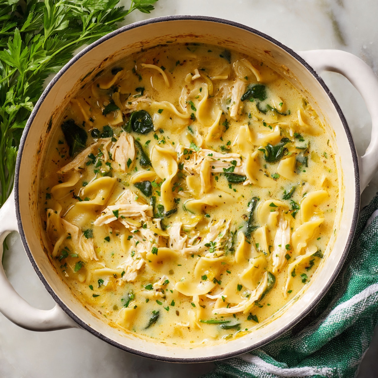 A white bowl filled with creamy soup containing three layers of ingredients: the base layer is light cream broth with specks of herbs, the middle layer has wavy, yellow pasta pieces floating with shredded white chicken meat, and the top layer is garnished with green chopped herbs and small bits of spinach. Small red pieces, likely tomatoes, add touches of color. The bowl is on a white marbled surface, with some green leafy herbs placed nearby and slices of crusty bread on the left side. photo taken with an iphone --ar 4:5 --v 7