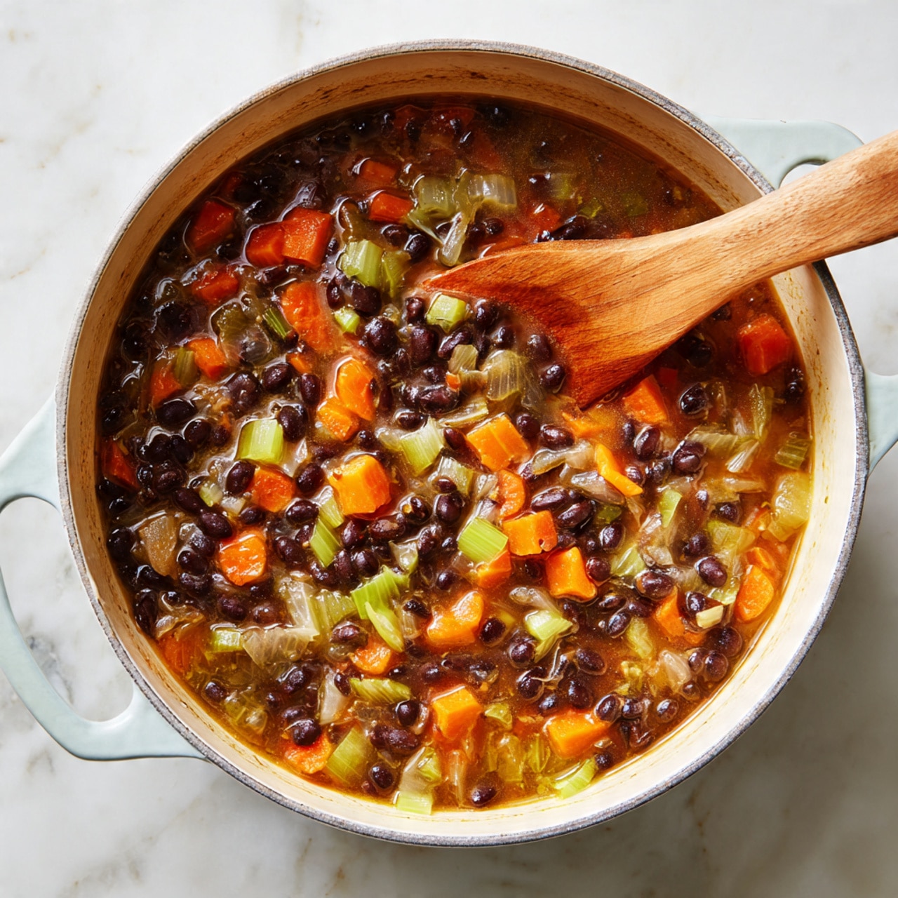 A close-up top view of a white pot filled with a thick vegetable and black bean stew, showing layers of dark black beans, orange carrot slices, pale green celery pieces, and translucent cooked onions mixed in a light brown broth. A wooden spoon is partly submerged on the right side, stirring the mixture, with the pot placed on a white marbled surface. The pot’s outer edge is slightly visible, showing a hint of light blue color. photo taken with an iphone --ar 4:5 --v 7