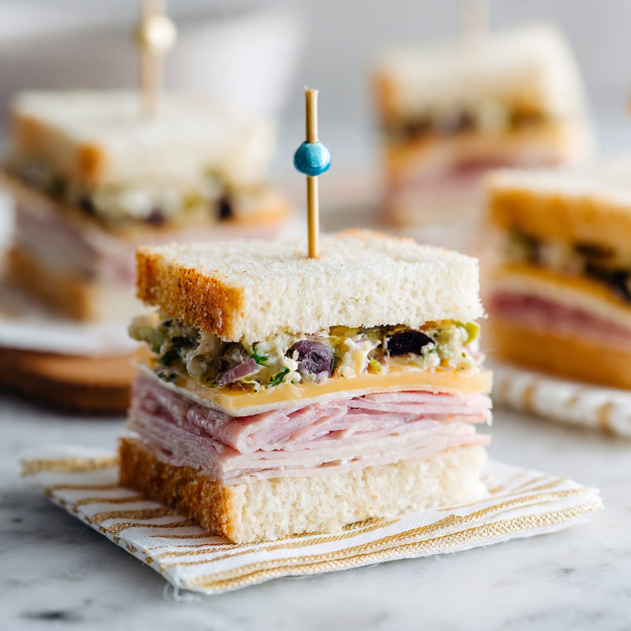 This image shows a sandwich cut into square pieces, each secured with a small wooden skewer topped with colorful beads in yellow, red, blue, and green. The sandwich has two thick layers of light brown, crusty bread with a soft, airy inside. Inside each piece are several thin layers of pink deli meat stacked neatly, with a layer of chopped vegetables including green, orange, and white bits placed on top of the meat. The sandwich pieces are arranged on a white marbled surface with some vegetable bits scattered around. Photo taken with an iphone --ar 4:5 --v 7
