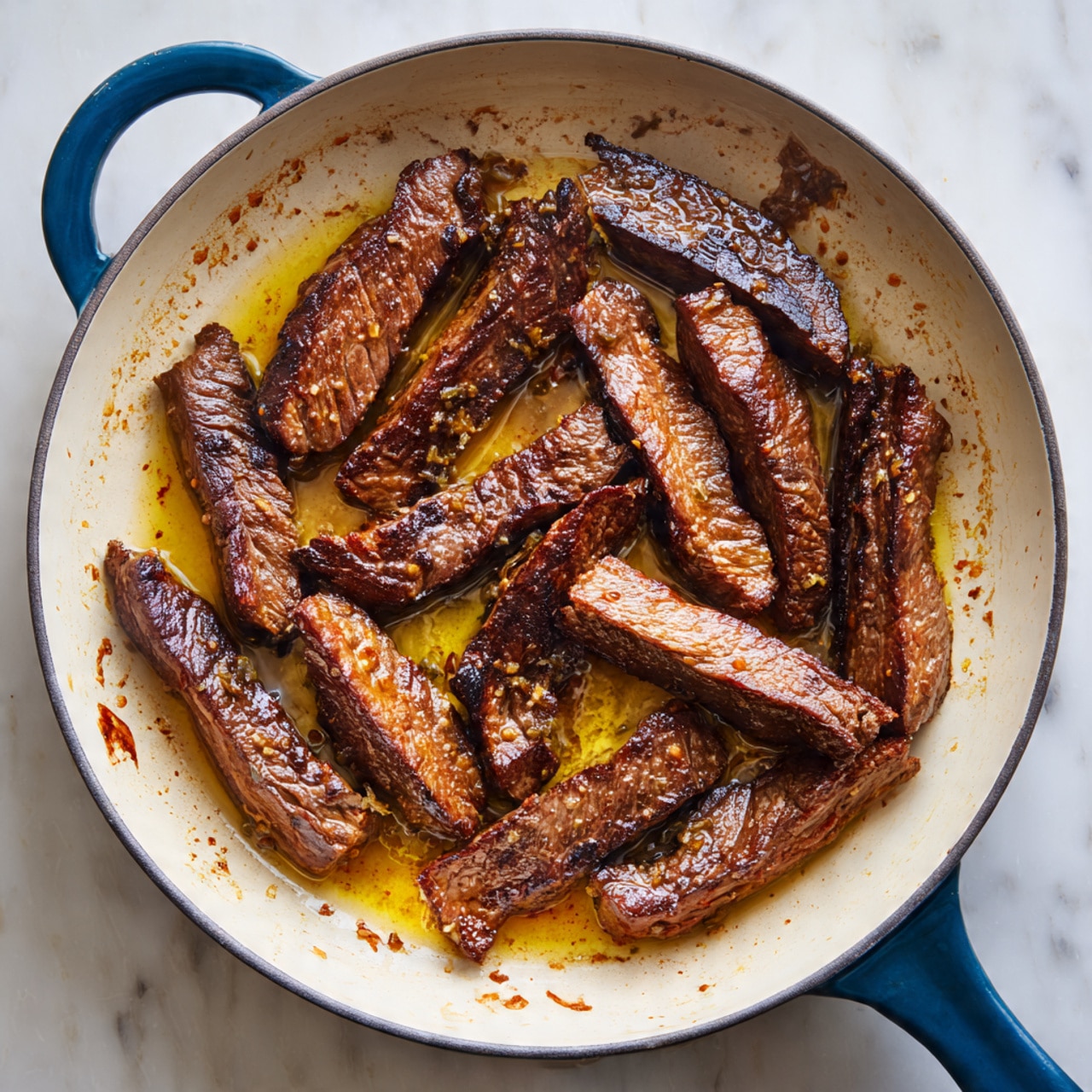 The image shows about fifteen dark brown strips of cooked meat with a shiny oily texture, spread out in an off-white pan with a light pattern of browned stuck bits on the bottom. The meat strips vary in size and shape, with some looking thicker and others thinner, lying flat with visible grill marks and a slightly rough surface. The pan has a thick blue handle visible on the lower right side, and it sits on a white marbled surface. There are small pools of golden oil around the meat, adding to the glossy appearance. Photo taken with an iphone --ar 4:5 --v 7