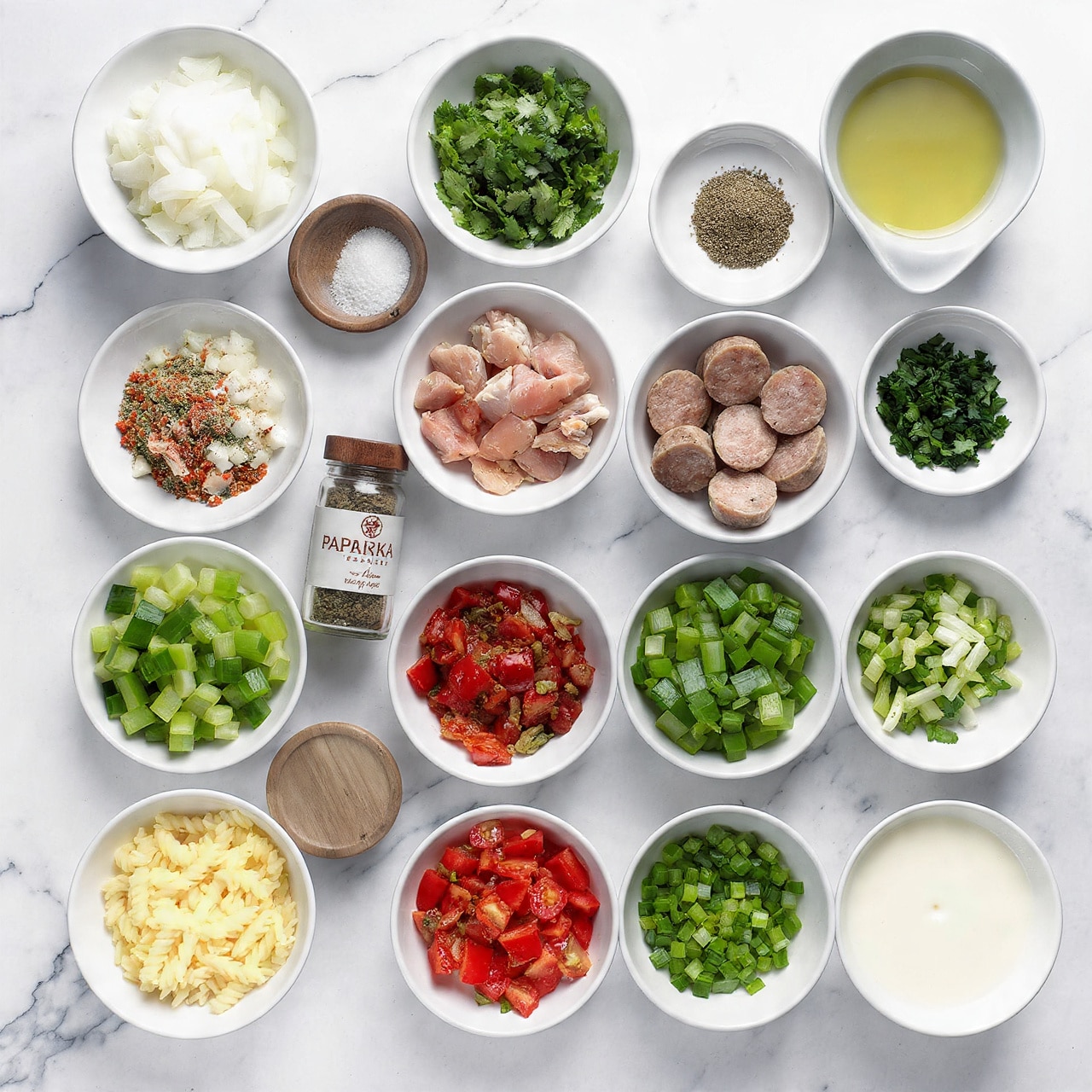 A top-view image showing various ingredients arranged neatly on a white marbled surface. There are 13 white bowls of different sizes containing chopped onions, fresh green cilantro leaves, vegetable oil, raw cut chicken pieces, sliced sausage, canned tomatoes, diced green bell peppers, chopped celery, chopped green onions, pasta, and milk or cream. Two small wooden bowls hold salt and black pepper. Two clear glass spice jars with wooden lids labeled