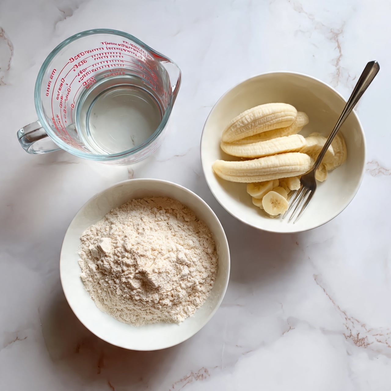 The image shows three items placed on a white marbled surface: a clear glass measuring cup filled with water marked in red measurements on the left, a white bowl with white flour inside in the front, and another white bowl to the right holding a peeled banana with part of it mashed and a silver fork resting on the bowl. The flour is loosely piled, light beige inside the bowl, the banana's texture contrasts between smooth and mashed parts, and the water is clear and reflective. Photo taken with an iphone --ar 4:5 --v 7