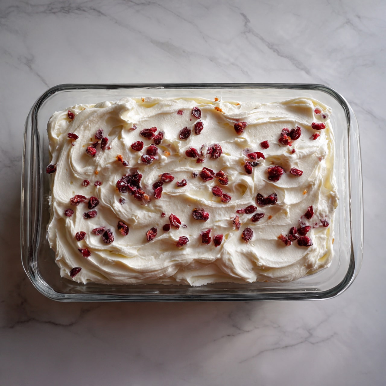A glass rectangular dish filled with a top layer of smooth white frosting that has soft swirls and waves on the surface. Scattered evenly across the frosting are small, dark red dried fruit pieces, adding a speckled texture and color contrast. The dish is set on a white marbled surface. photo taken with an iphone --ar 4:5 --v 7