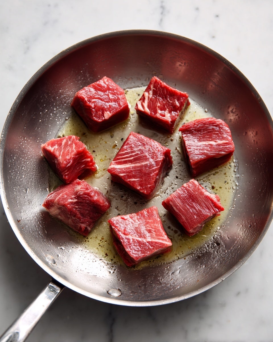 In a metal pan, there are nine pieces of raw meat, each square and thick, with a rich red color and some white fat veins. The pieces are spread out slightly, cooking in a light layer of shiny oil that covers the bottom of the pan. The pan has a metallic silver surface inside, slightly worn from use. The scene is set on a white marbled surface. photo taken with an iphone --ar 4:5 --v 7