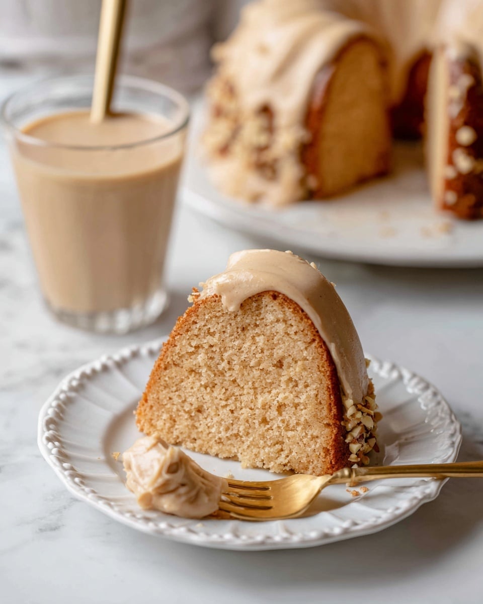 A single slice of light brown cake with a soft, spongy texture sits on a white plate with decorative edges, topped with a smooth, light beige frosting layer that also wraps the cake’s side, sprinkled with small nut pieces near the bottom edge. A gold fork is digging into the slice from the front left, held by a woman's hand that is slightly visible at the edge. Behind the plate is a clear glass filled with a creamy light yellow beverage and a straw, placed on a white marbled surface. In the blurred background, a larger bundt-style cake with the same frosting sits on a white plate with a gold rim. Photo taken with an iphone --ar 4:5 --v 7