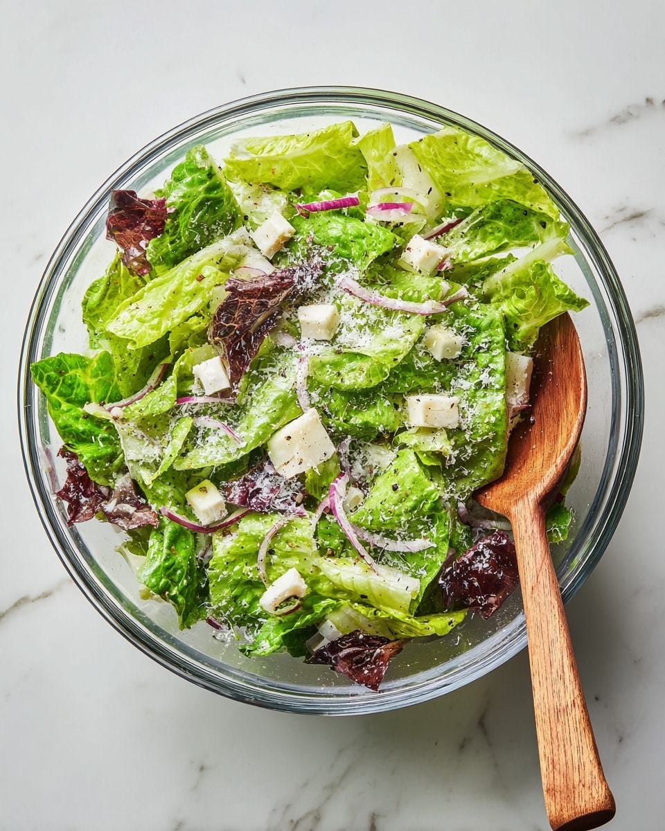 A clear glass bowl filled with a fresh salad placed on a white marbled surface. The salad has one main layer of bright green romaine lettuce leaves with a mix of darker leafy greens underneath. Scattered on top are small white cubes of cheese and thin slices of purple-red onion. The salad is sprinkled with finely grated white cheese and a light dusting of black pepper. A wooden spoon rests on the right side inside the bowl, partially submerged in the salad. Photo taken with an iphone --ar 4:5 --v 7
