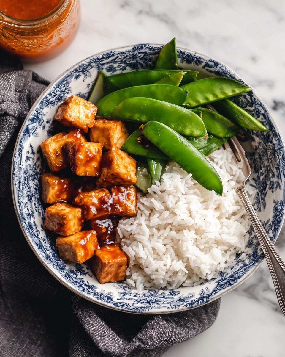 A white bowl with a blue pattern holds a layer of white rice on the bottom right, next to bright green snow peas on the upper right sitting directly on the rice, and golden brown cubes of tofu coated in a shiny, slightly dark sauce placed on the left side. A silver fork rests in the rice on the bottom right inside the bowl. The bowl is on top of a dark gray cloth, all set on a white marbled surface. A jar with orange sauce is seen on the top left outside the bowl. photo taken with an iphone --ar 4:5 --v 7