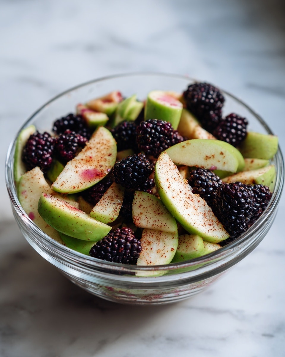 A clear glass bowl is filled with a mix of sliced green apples and whole blackberries, all coated in a light, reddish spice or seasoning. The apple slices are layered unevenly throughout, showing smooth, pale green skin with some pinkish spice marks, while the plump blackberries add a textured, deep black-purple contrast scattered around and on top. The bowl sits on a white marbled surface, with soft natural light highlighting the fresh and colorful fruit mix. photo taken with an iphone --ar 4:5 --v 7