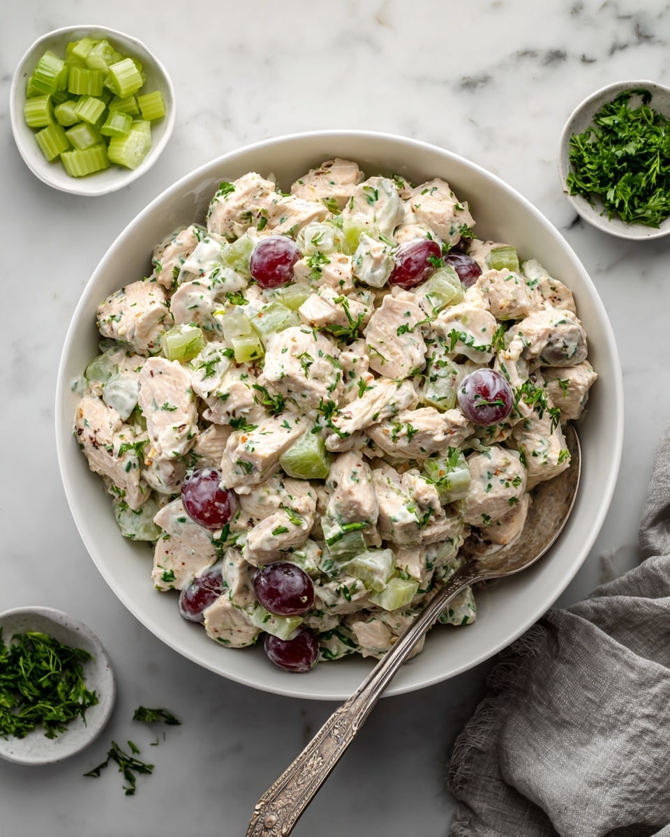 A clear glass bowl holds a creamy chicken salad mixed evenly with chopped light beige chicken pieces, dark red and purple grape slices, and bright green chopped celery and green onions. Thin almond slices add a light toasted brown texture. The creamy dressing coats the ingredients, giving the salad a smooth, slightly shiny look. A silver spoon rests inside the bowl, and the bowl is placed on a white marbled surface photo taken with an iphone --ar 4:5 --v 7