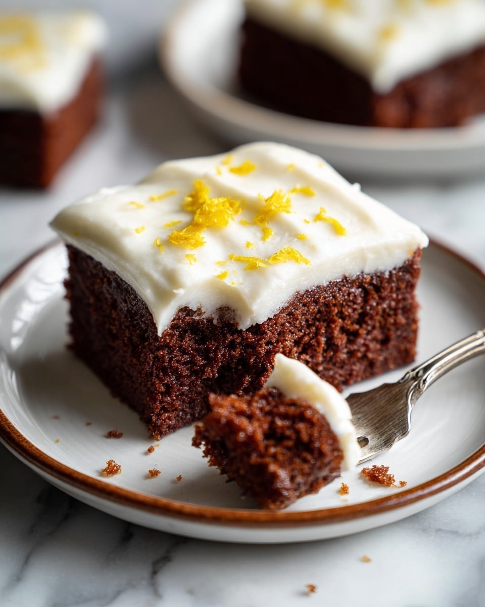 A square dark brown chocolate cake lies on crumpled parchment paper, topped with one thick creamy white frosting layer that is spread unevenly to the edges. Bright yellow lemon zest is scattered lightly over the frosting, adding vibrant specks of color. To the left of the cake, two halves of a fresh yellow lemon sit on the white marbled surface. In the upper right corner, a white plate with a thin brown rim holds silver forks, resting on the same white marbled surface. Photo taken with an iphone --ar 4:5 --v 7