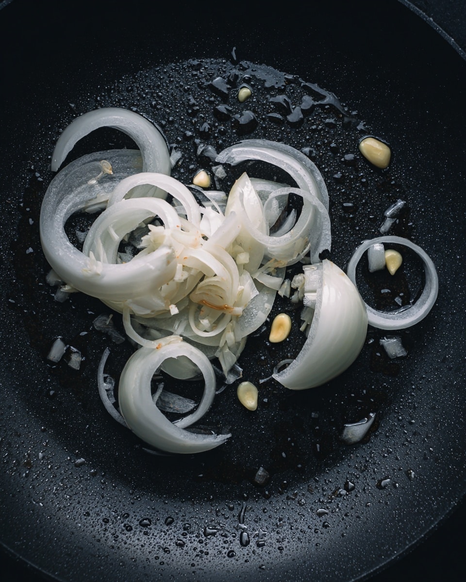 A black frying pan with a few slices of white onion and whole small pieces of peeled yellow garlic cloves spread out inside, surrounded by small droplets of cooking oil that glisten under the light; the onions are mostly curved and translucent with smooth surfaces, arranged in a loose cluster around the center of the pan, with the garlic pieces nestled between them, all on a dark surface with a slightly shiny texture. photo taken with an iphone --ar 4:5 --v 7