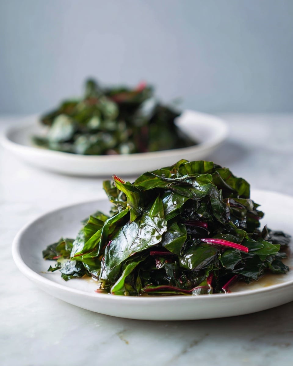 Two white plates sit on a white marbled surface, each holding a small pile of cooked leafy greens. The greens are dark green with some red stems mixed in, looking tender and shiny from cooking. The greens are layered loosely and show some light texture from wilting, with small pieces scattered slightly beyond the main pile. The lighting highlights the freshness and moisture of the greens. photo taken with an iphone --ar 4:5 --v 7