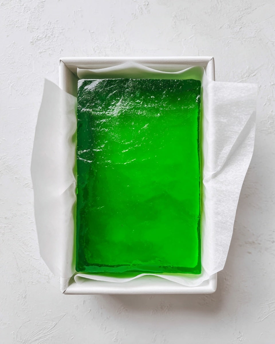 A single thick rectangular layer of bright green jelly sits inside a white parchment paper-lined tray. The jelly is smooth with a shiny, slightly rippled surface, filling most of the tray with edges softly touching the white paper. The tray rests on a white marbled texture surface. photo taken with an iphone --ar 4:5 --v 7