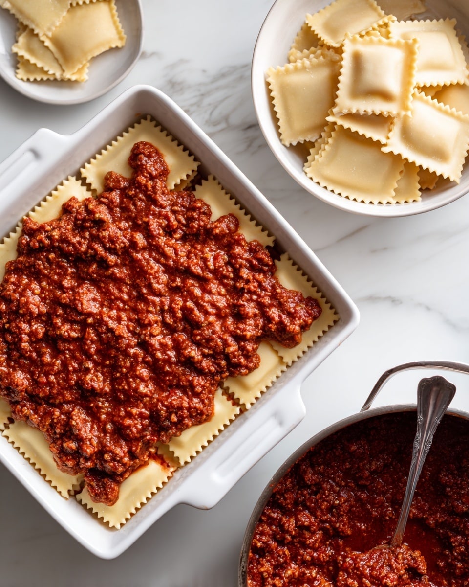 The image shows a white rectangular baking dish filled with the first layer of lasagna. The bottom layer is made of a thick, chunky red meat and tomato sauce spread evenly across the base. On top of the sauce, there is a single layer of uncooked square ravioli with wavy edges, arranged closely but not overlapping. Next to the baking dish, there is a white bowl filled with more uncooked ravioli, and a dark pan containing more red meat sauce with a metal spoon resting in it. The entire setup is placed on a white marbled surface. Photo taken with an iphone --ar 4:5 --v 7