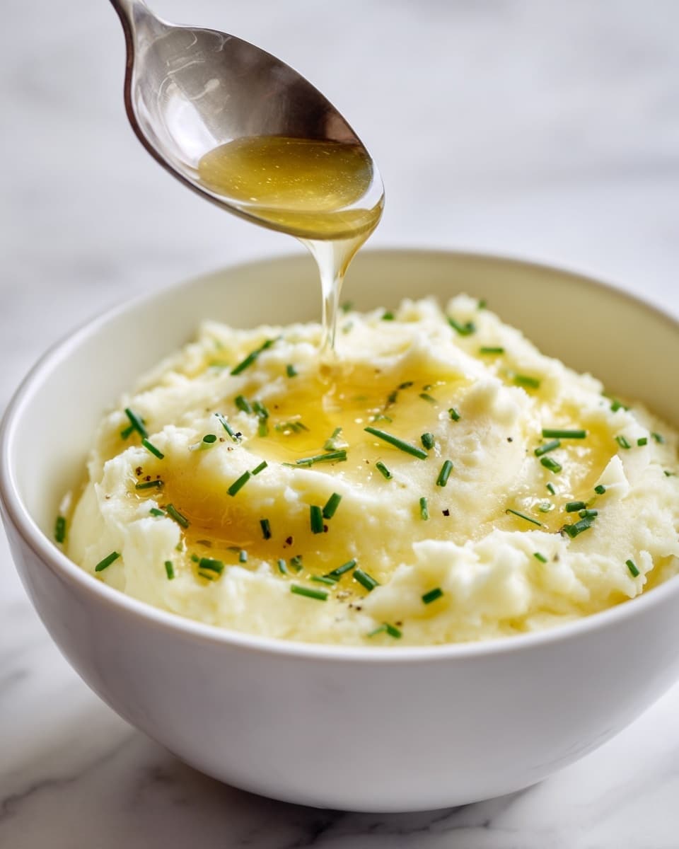 A white bowl holds a creamy layer of pale yellow mashed potatoes with a smooth texture, topped with small green chive pieces scattered around. A spoon above the bowl drizzles a shiny, golden melted butter liquid that pools gently on the surface, adding a rich gloss. The background is a soft white marbled surface, creating a bright and fresh look. Photo taken with an iphone --ar 4:5 --v 7