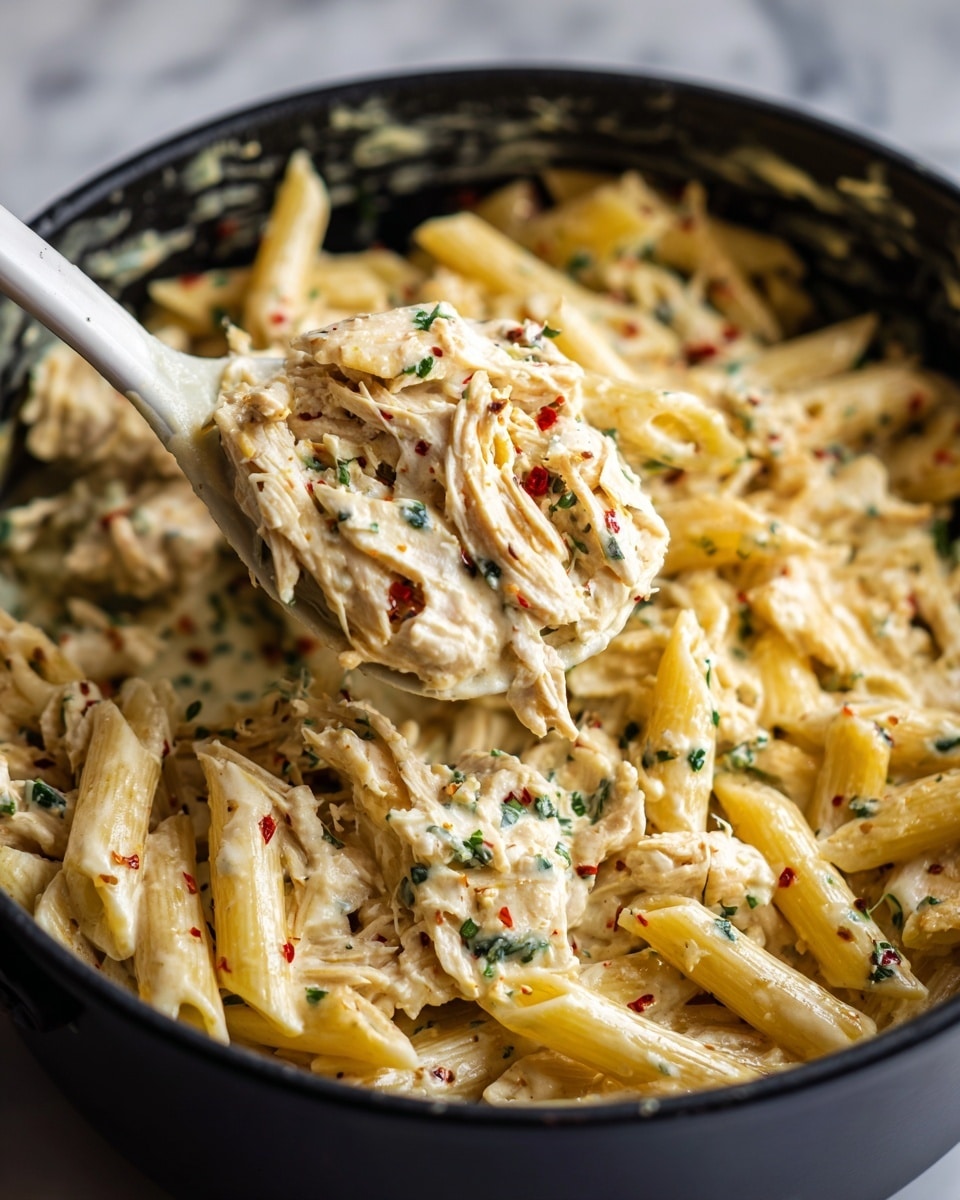 A close-up view of a creamy pasta dish in a black pot. The dish has two main layers: pale yellow penne pasta scattered throughout and shredded white chicken mixed in thick creamy sauce with small red and green herb bits visible. A white spoon lifts a portion, showing the mix of pasta and chicken coated evenly with the sauce. The pot's inside looks slightly glossy, holding the warm and rich pasta mix. The background is a white marbled texture. Photo taken with an iphone --ar 4:5 --v 7