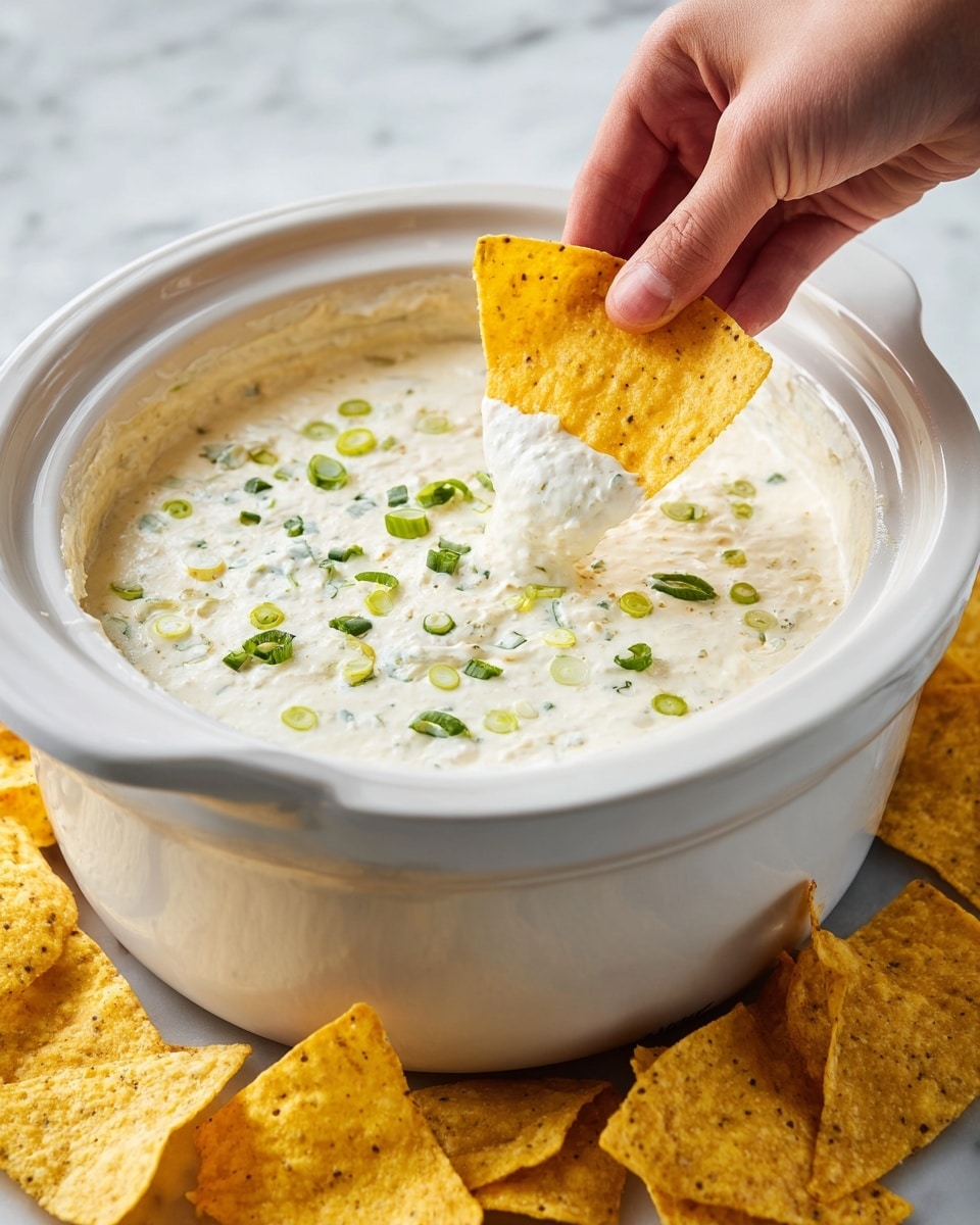 A white crockpot filled with creamy white cheese dip topped with small green onion slices floating on the surface; a woman's hand dips a yellow tortilla chip with a crisp texture into the smooth cheese, showing the chip partially covered in the cheese dip; several more yellow tortilla chips with a rough texture lay scattered around the base of the crockpot on a white marbled surface photo taken with an iphone --ar 4:5 --v 7
