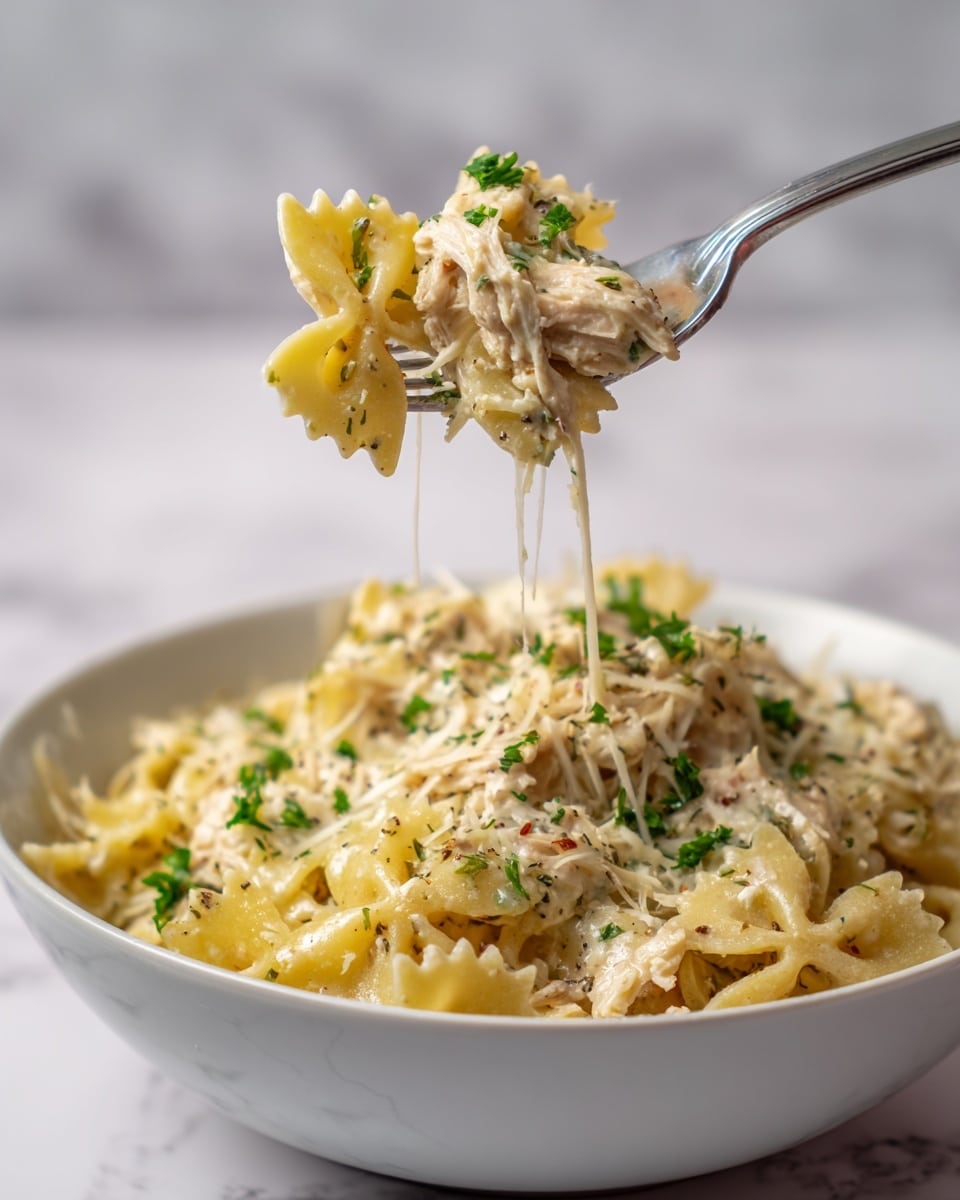 The image shows a close-up of a silver fork lifting a small bundle of pasta from a white bowl filled with creamy pasta. The pasta is layered with yellow bow-tie shapes, shredded chicken coated in a creamy white sauce with herbed specks, and thin strands of melted cheese, with some fresh green parsley bits scattered on top. The white bowl beneath holds a large serving of the same creamy pasta with visible herbs and chicken pieces, all set on a white marbled surface with a plain light background. photo taken with an iphone --ar 4:5 --v 7