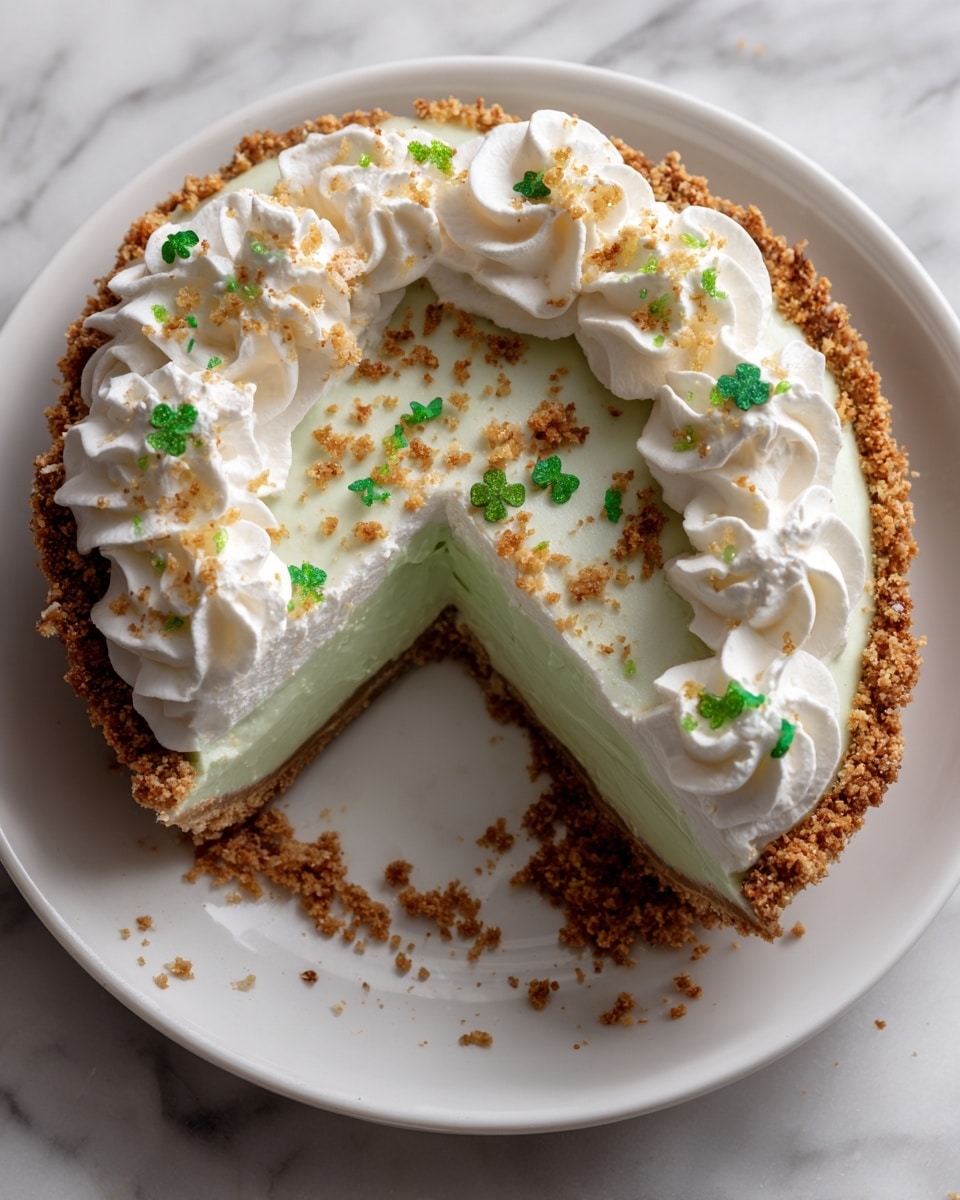 A pie sits on a white plate with a crumbly golden-brown crust at the bottom and edges. The middle layer is light green, smooth, and creamy. On top, a thick layer of white whipped cream is spread evenly, with swirls piped around the edge. Small green clover-shaped sprinkles are scattered on the whipped cream and a few are on the plate. The plate is on a white marbled surface. Photo taken with an iphone --ar 4:5 --v 7