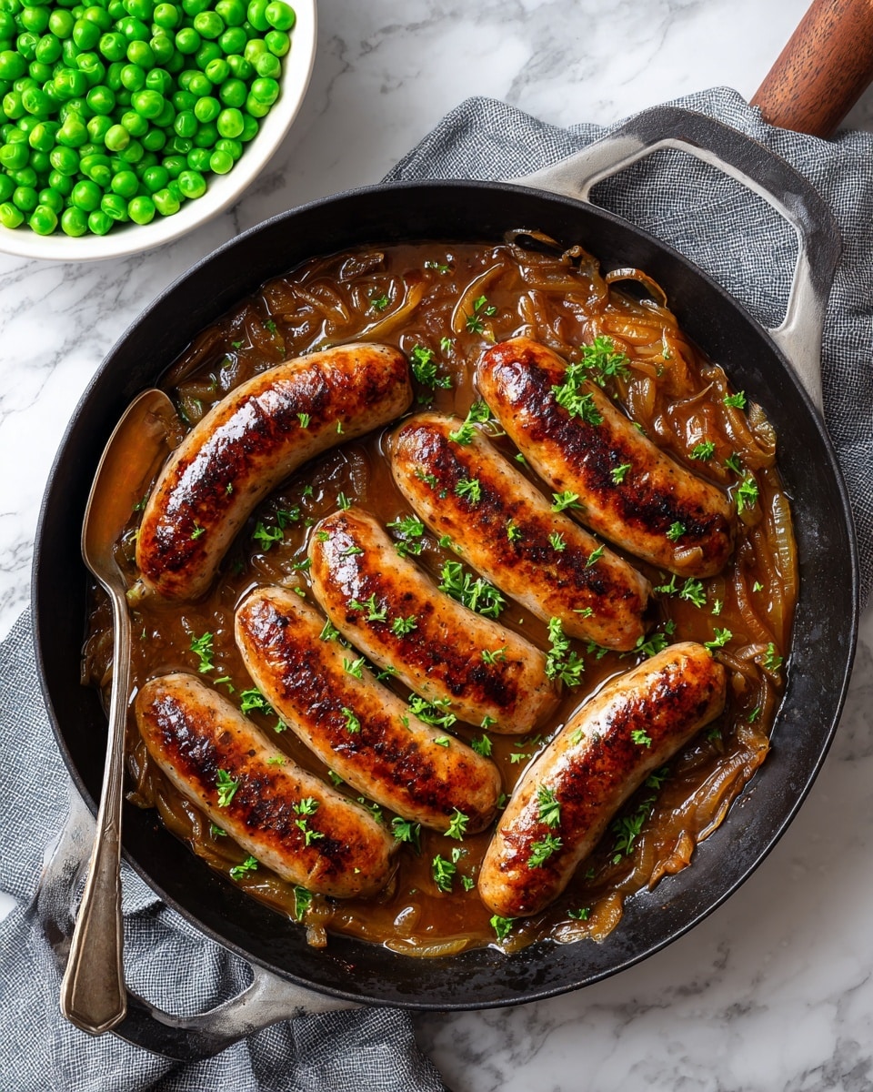 A black cast iron pan filled with six browned sausages arranged in two rows, sitting on a thick brown sauce mixed with cooked onions. The sausages have a shiny surface with some char marks, and the sauce is garnished with small green parsley leaves scattered evenly across the dish. A metal spoon rests in the pan, partially submerged in the sauce near the bottom left. Next to the pan, there is a white bowl filled with bright green peas on a folded gray cloth, all placed on a white marbled surface. Photo taken with an iphone --ar 4:5 --v 7