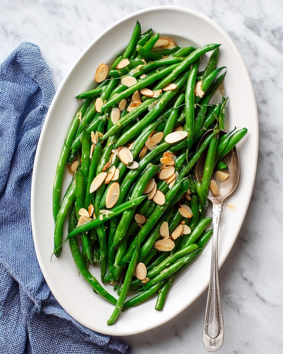 Inside a black frying pan with a silver handle, there is one thick layer of bright green green beans mixed with small pieces of light brown toasted garlic and tiny bits of seasoning scattered throughout. The green beans have a shiny, slightly oily texture showing they were cooked. The pan rests on a folded white cloth with thin dark blue stripes, placed on a white marbled surface. Photo taken with an iphone --ar 4:5 --v 7