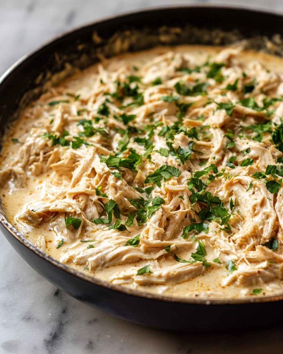 A close-up view of creamy shredded chicken in a black skillet, with a light beige sauce that looks smooth and thick, showing visible shredded chicken pieces mixed evenly. Scattered on top are small pieces of fresh green herbs. The skillet sits on a white marbled surface. photo taken with an iphone --ar 4:5 --v 7