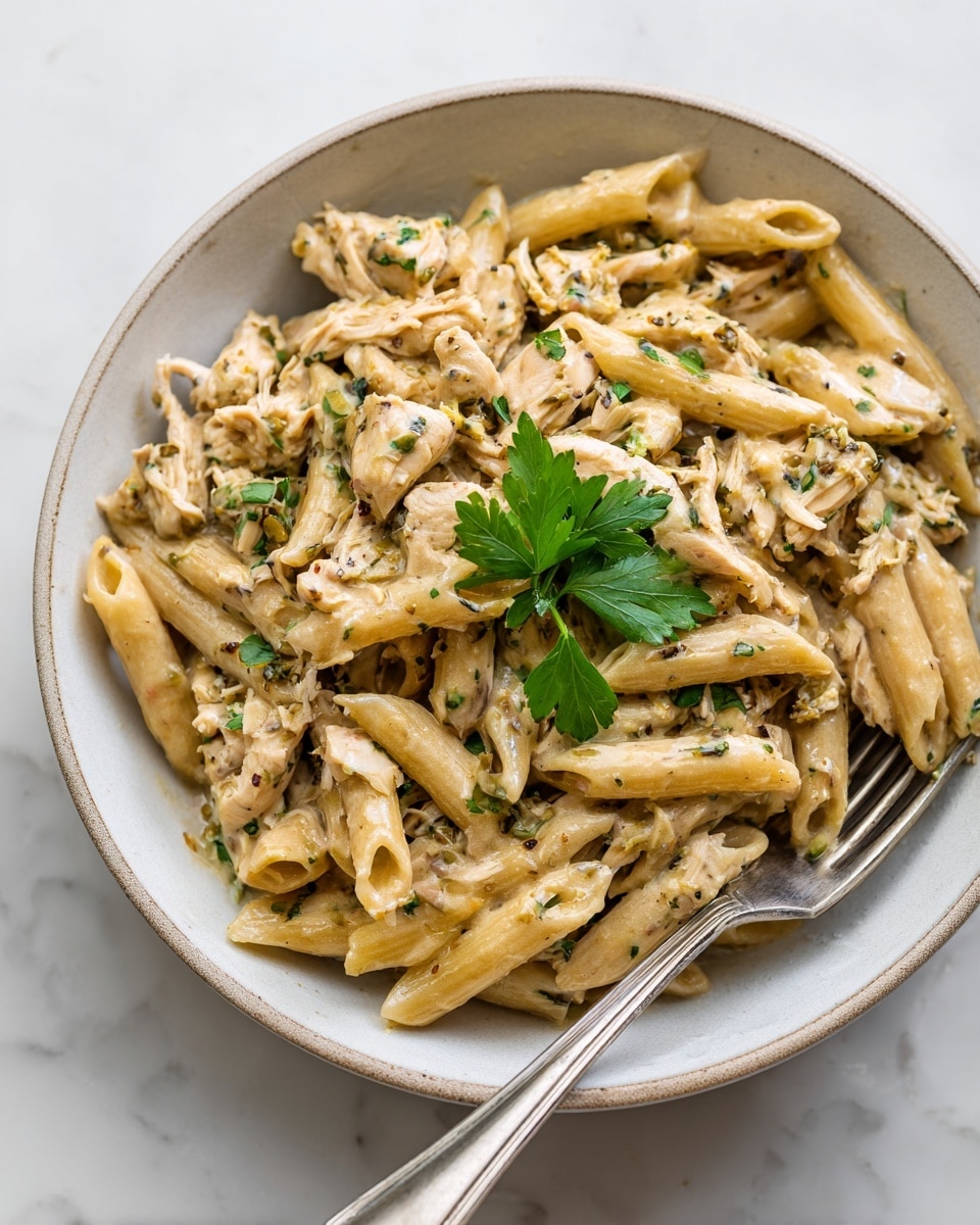 A close-up of a bowl filled with a creamy pasta dish featuring penne mixed with shredded chicken in a light beige sauce. The pasta and chicken are coated evenly, showing a slightly grainy texture with small green herb pieces scattered throughout. A small sprig of fresh parsley rests on top at the center, adding contrast with its deep green color. A fork is placed in the bowl on the right side, partially inserted into the pasta. The bowl is white with a rustic edge and sits on a white marbled surface. Photo taken with an iphone --ar 4:5 --v 7