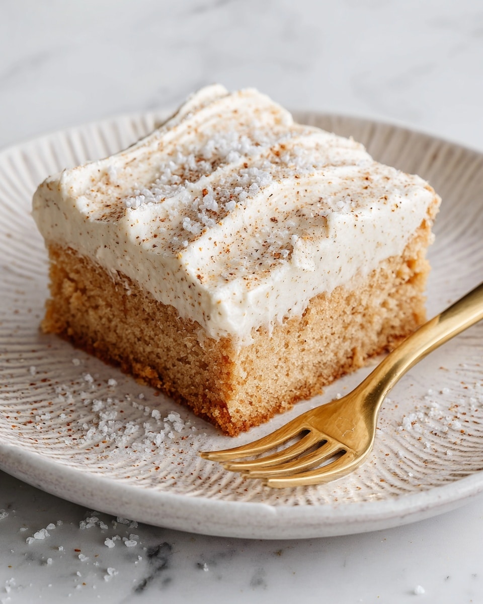 The image shows a single square piece of cake on a white plate with a textured pattern. The cake has two visible layers: the bottom layer is a light brown color with a soft texture, and the top layer is a thick, creamy white frosting sprinkled with fine brown specks and a dusting of white sugar crystals. The frosting has soft ridges running diagonally across its surface. Next to the cake on the plate is a shiny gold fork. The plate rests on a white marbled surface. photo taken with an iphone --ar 4:5 --v 7