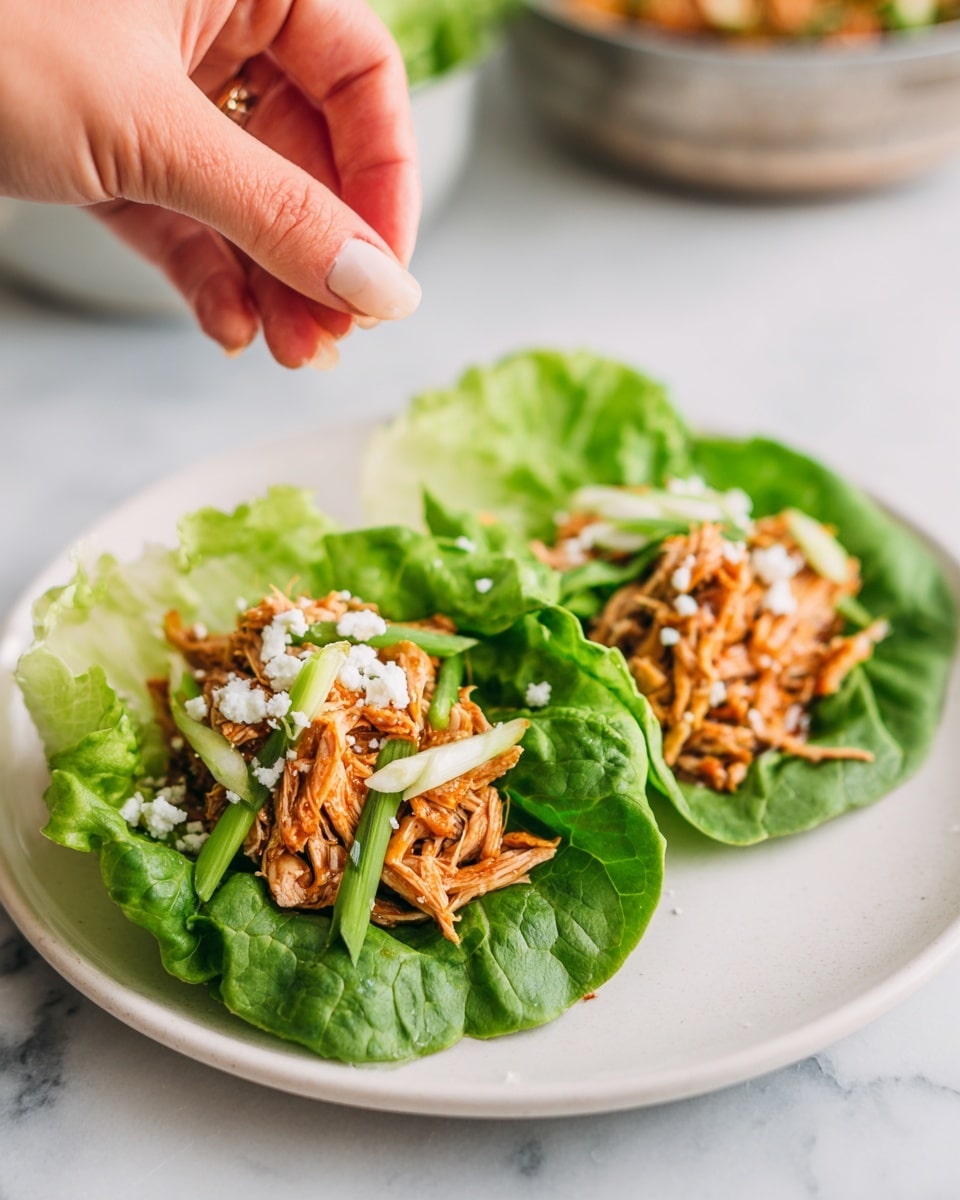 Two lettuce wraps sit on a white plate with crisp green leaves forming the base layer. On top of each leaf are pieces of orange-brown shredded meat mixed with slices of light green celery. A woman's hand is sprinkling small white crumbles over the meat and celery on one wrap. In the background, there is a blurry metal bowl filled with more shredded meat, resting on a white marbled texture. photo taken with an iphone --ar 4:5 --v 7