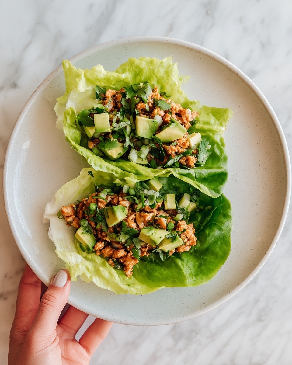 The image shows two lettuce cups on a white plate, each filled with shredded light brown meat and topped with a mix of green leafy herbs and small cubes of avocado. The lettuce leaves are bright light green with a fresh, crisp texture, wrapping the fillings neatly. There is a woman's hand holding one of the lettuce cups on the right side of the image. The background is a white marbled surface, giving a clean and fresh look to the dish. photo taken with an iphone --ar 4:5 --v 7