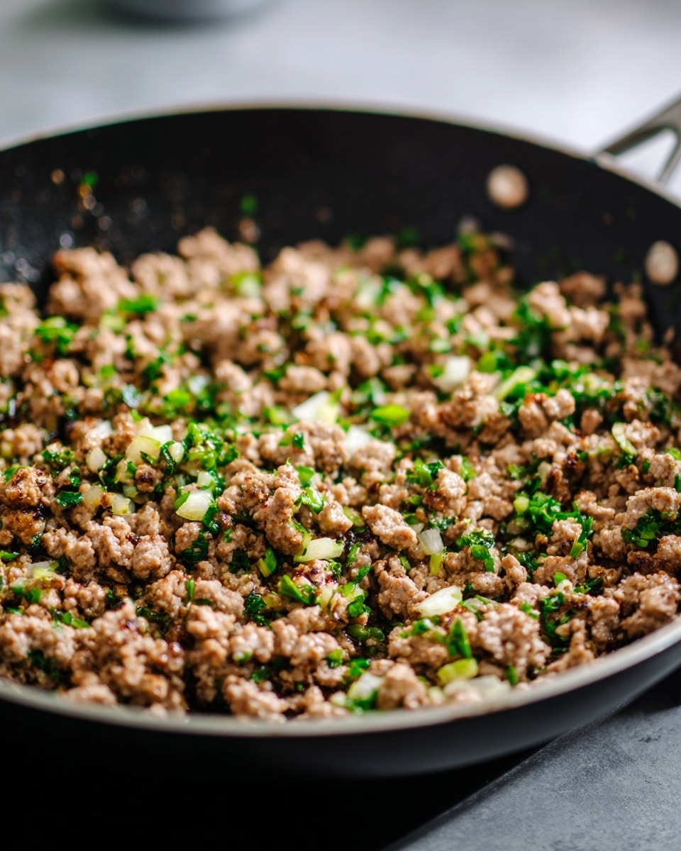 A close-up view of a large black skillet filled with browned ground meat mixed with small bits of green herbs and onions, sitting on a black stovetop. The meat is crumbly and cooked evenly, with tiny green pieces scattered throughout, showing a mix of cooked textures and slight moisture. The background is blurred but shows a grayish kitchen setting. photo taken with an iphone --ar 4:5 --v 7