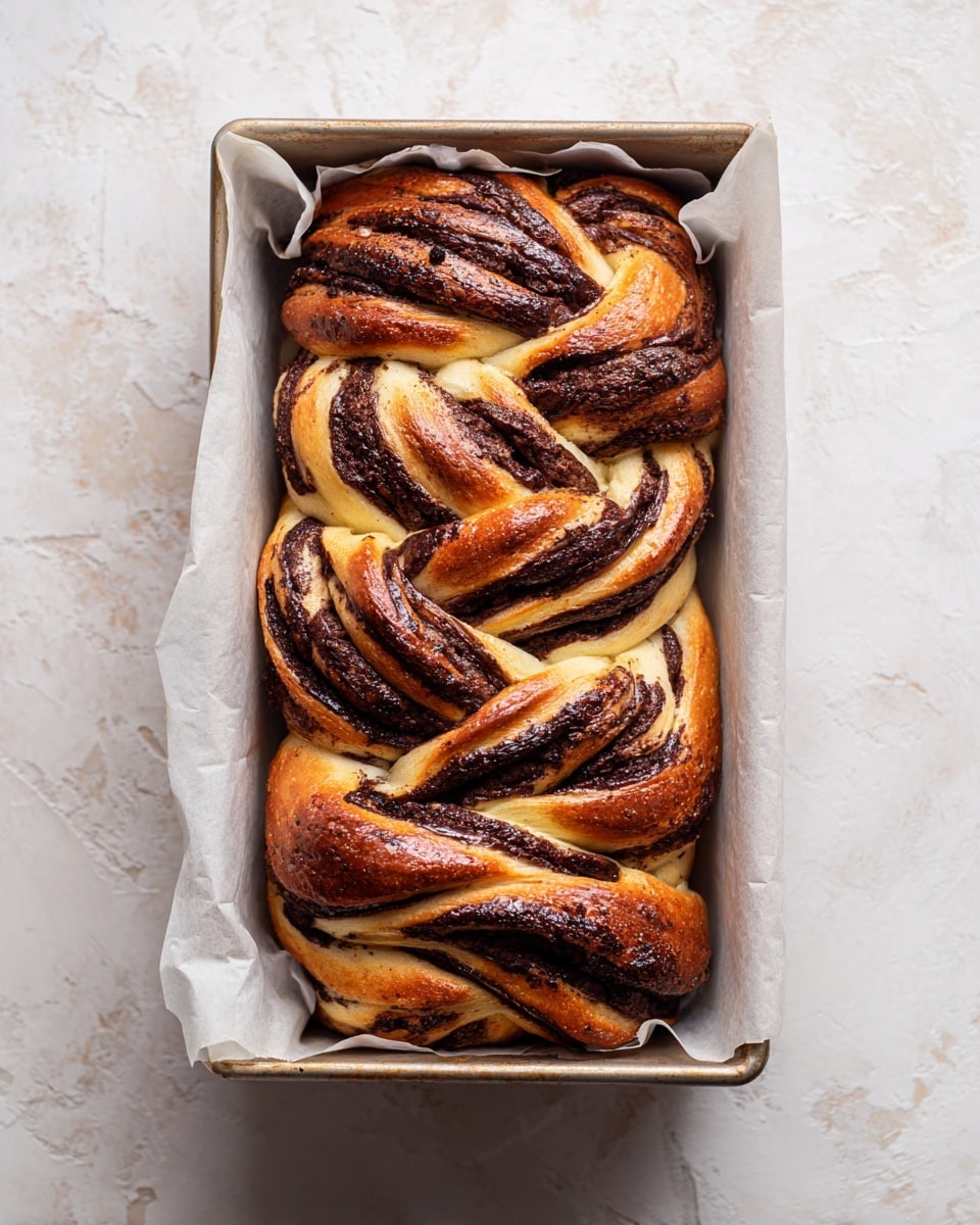 A freshly baked loaf of twisted bread sits inside a metal rectangular baking pan lined with white parchment paper. The bread has multiple thick layers woven together, showcasing a golden brown crust with darker chocolate-filled swirls running through the soft, light tan dough. The surface of the bread looks shiny and slightly crispy with rich dark brown patches from the chocolate, creating a marbled effect. The baking pan rests on a white marbled textured surface. photo taken with an iphone --ar 4:5 --v 7