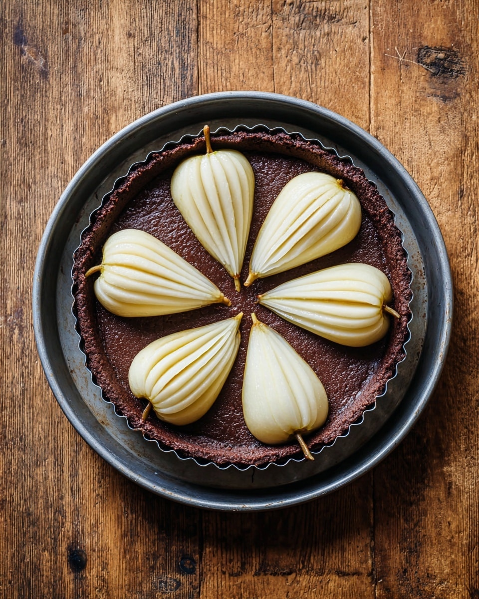 A round tart pan with a fluted edge holds a thick, dark brown chocolate batter spread evenly as the base layer. On top, six peeled pear halves are arranged in a circular pattern with the wider ends facing outward, their light cream color contrasting against the dark batter. The pears have thin vertical ridges creating a subtle striped texture. The pan sits centered on a silver baking tray with slight wear marks, placed on a wooden surface. photo taken with an iphone --ar 4:5 --v 7